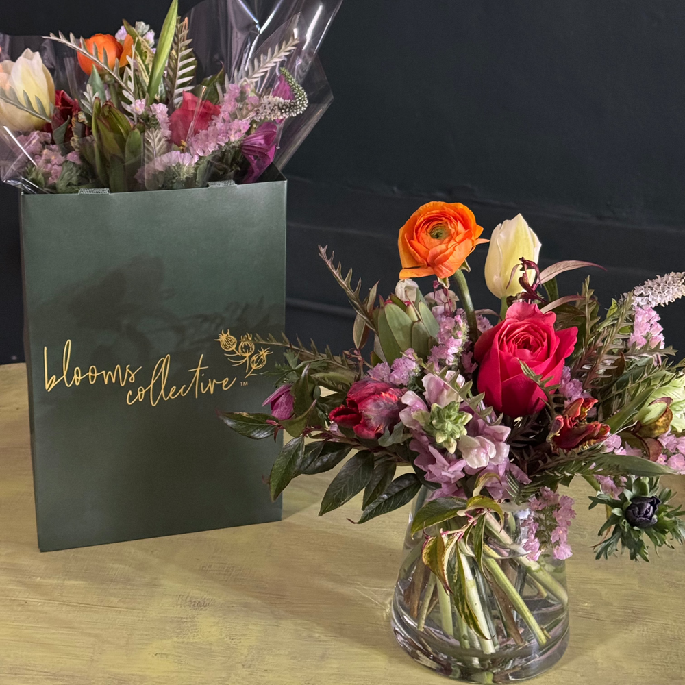 A hand-tied bouquet of fresh flowers featuring various colors like red, orange, and pink, presented in a clear glass vase with a green gift bag in the background.