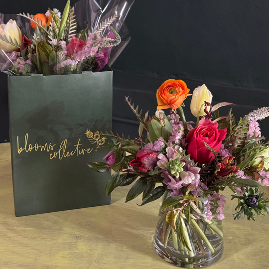 A hand-tied bouquet of fresh flowers featuring various colors like red, orange, and pink, presented in a clear glass vase with a green gift bag in the background.