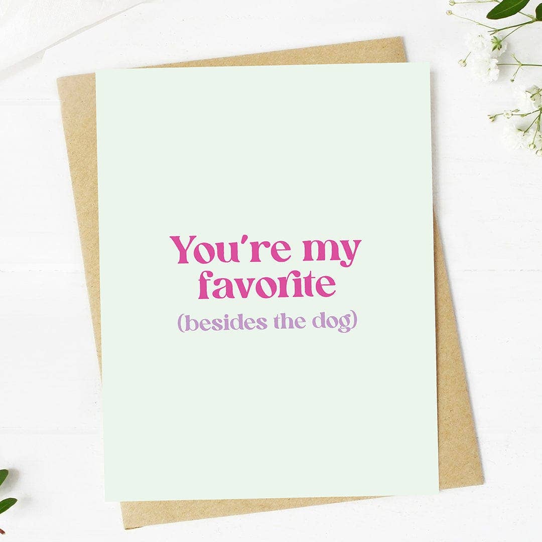 Greeting card with the text 'You're my favorite (besides the dog)' printed in pink on a light background, accompanied by a kraft envelope.
