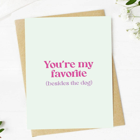Greeting card with the text 'You're my favorite (besides the dog)' printed in pink on a light background, accompanied by a kraft envelope.