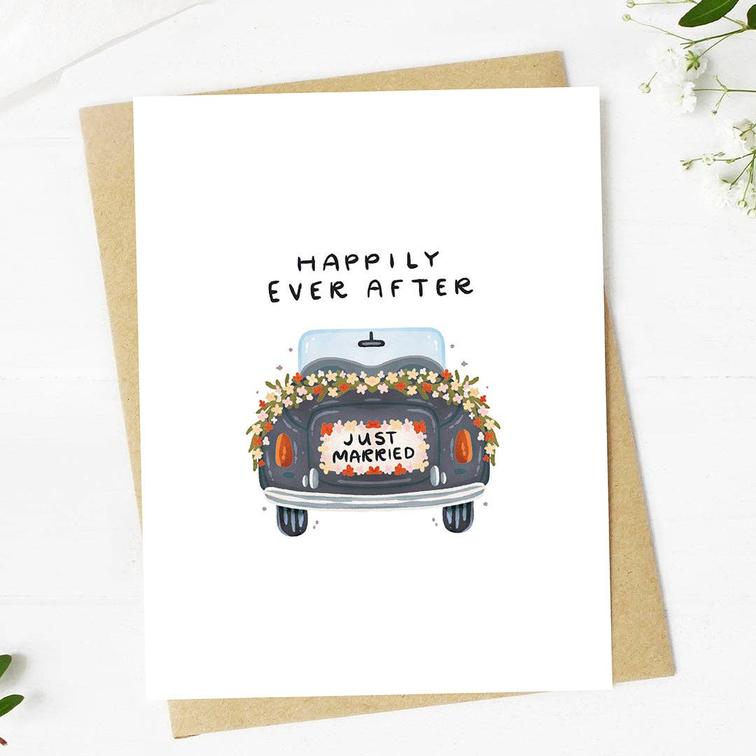 A greeting card with a hand-drawn illustration of a car with a 'Just Married' sign and flowers on the back, with the text 'HAPPILY EVER AFTER' above it. The card is placed on a white background with a kraft envelope beside it.