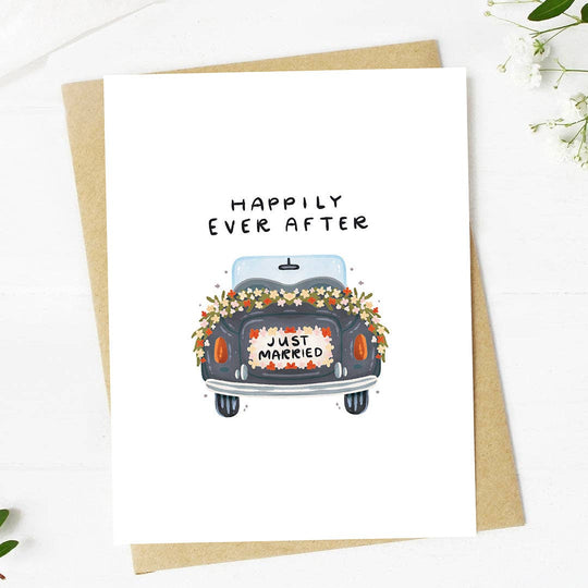 A greeting card with a hand-drawn illustration of a car with a 'Just Married' sign and flowers on the back, with the text 'HAPPILY EVER AFTER' above it. The card is placed on a white background with a kraft envelope beside it.