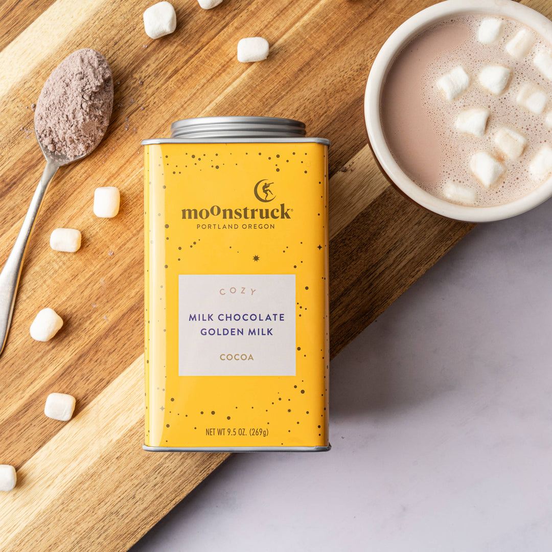 A tin of Cozy: Golden Milk Chocolate Hot Cocoa with ginger and turmeric, displayed next to a spoon and a cup of hot cocoa with marshmallows on a wooden surface.