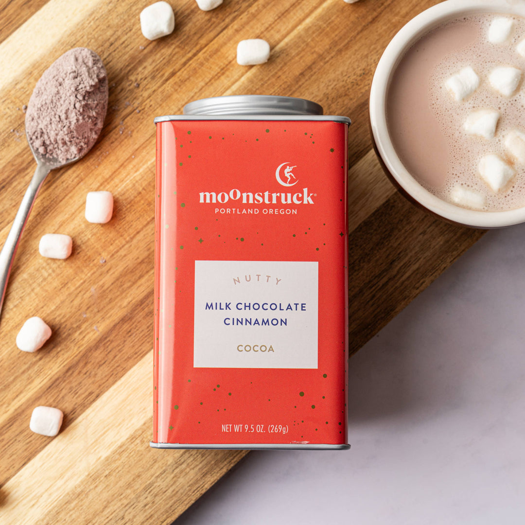 A tin of Moonstruck Milk Chocolate Hot Cocoa with cinnamon on a wooden surface, alongside a spoon with cocoa on it and a bowl of hot cocoa with marshmallows.