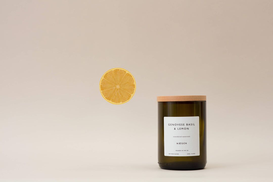 A green glass wine bottle style candle with a wooden lid, labeled 'Genovese Basil & Lemon', with a slice of lemon floating above it against a beige background.