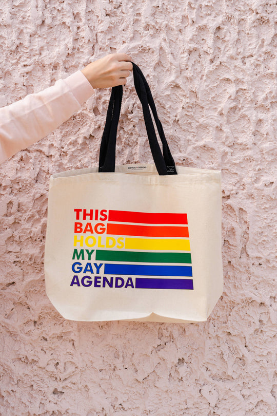 Tote bag with rainbow design and text held against a textured wall.