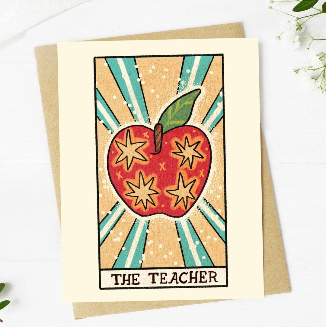 A tarot-themed greeting card with a hand-drawn illustration of a red apple with stars and the word 'THE TEACHER' below it. The card is accompanied by a beige envelope and is placed on a white background with some floral design at the bottom.