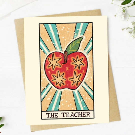 A tarot-themed greeting card with a hand-drawn illustration of a red apple with stars and the word 'THE TEACHER' below it. The card is accompanied by a beige envelope and is placed on a white background with some floral design at the bottom.