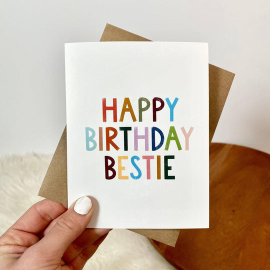 A hand holding a birthday card with 'HAPPY BIRTHDAY BESTIE' written in colorful letters. The card is accompanied by a brown envelope.