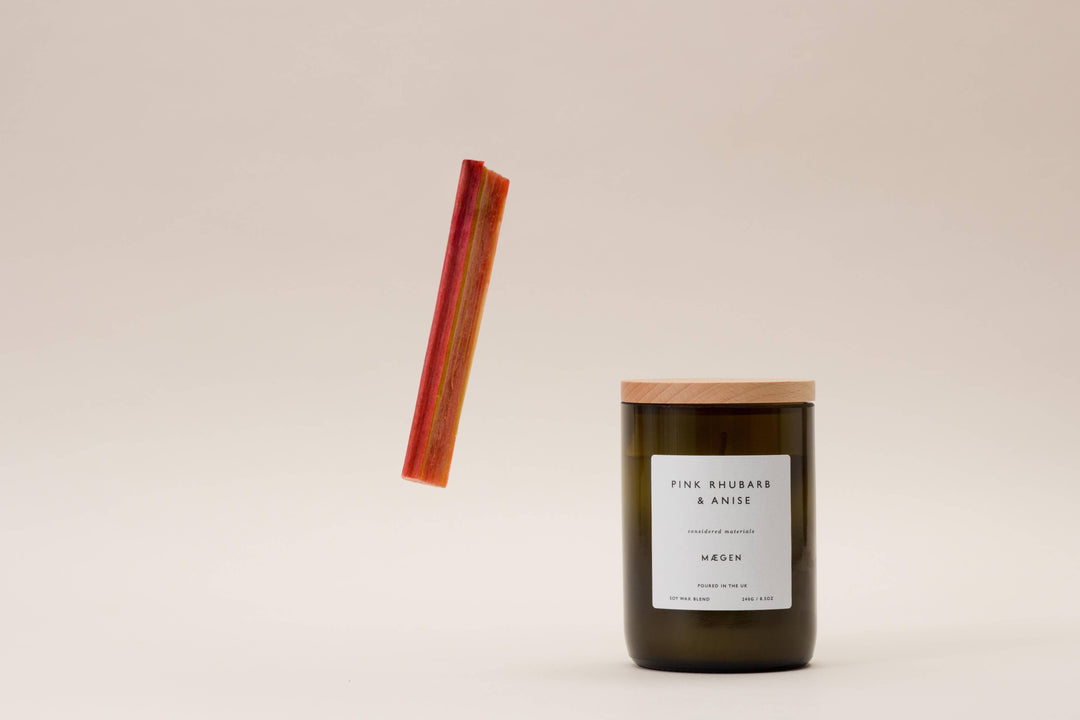 A wine bottle style candle with a wooden lid, green glass, and a pink rhubarb anise label, accompanied by a floating pink rhubarb stick.