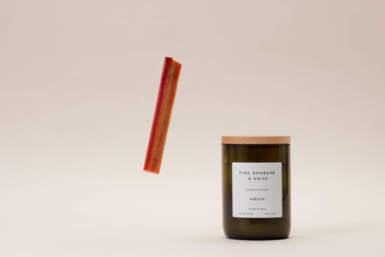 A wine bottle style candle with a wooden lid, green glass, and a pink rhubarb anise label, accompanied by a floating pink rhubarb stick.