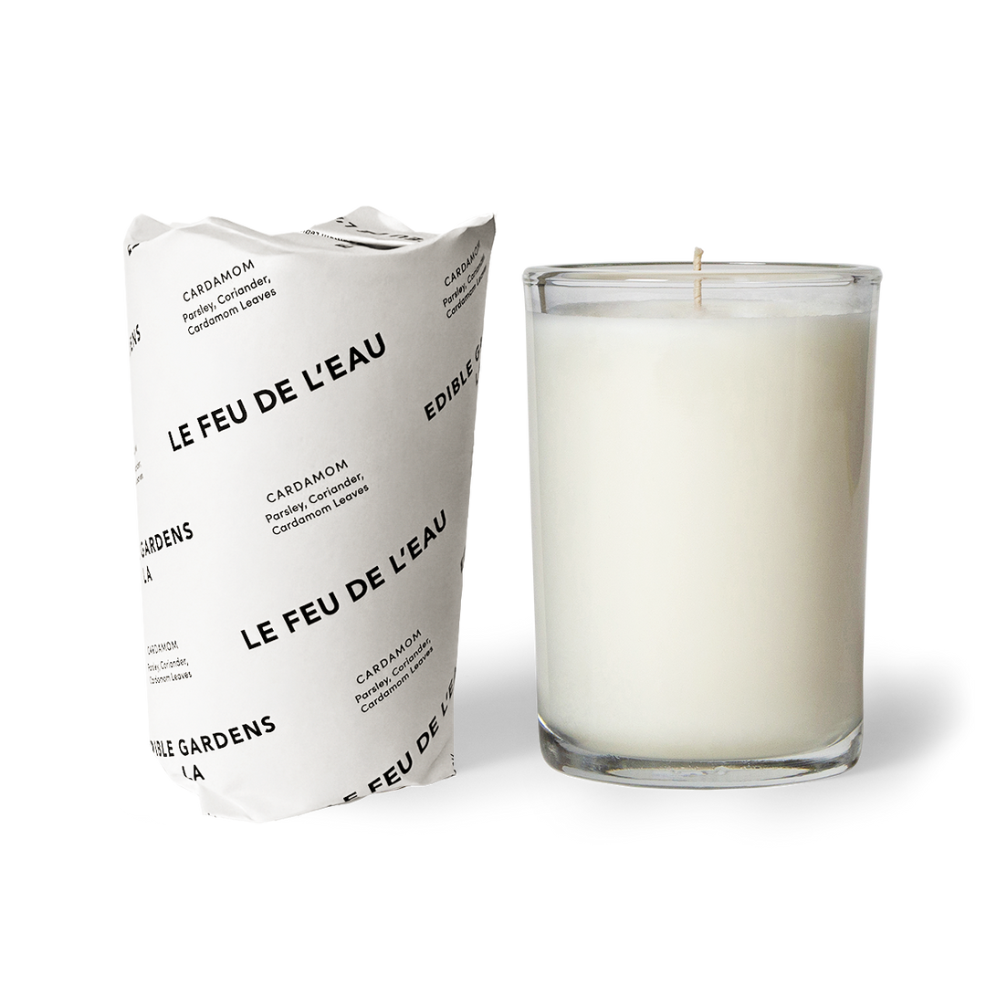 A white candle in a clear glass container, with a label wrapped around the paper packaging reading 'LE FEU DE L'EAU' and 'CARDAMOME'.