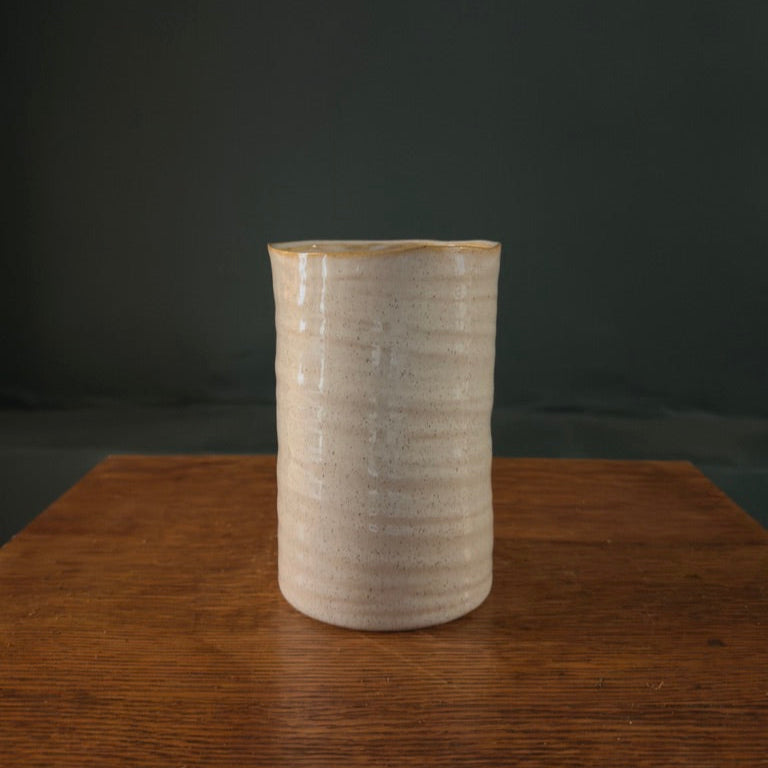 A beige ceramic vase with vertical ridges, displayed on a wooden surface against a dark background.