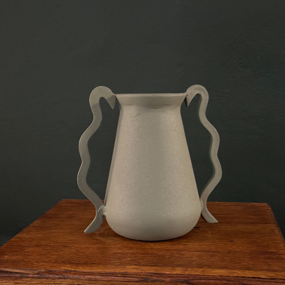 A sage green aluminum vase with double wavy handles, displayed on a wooden surface against a dark background.