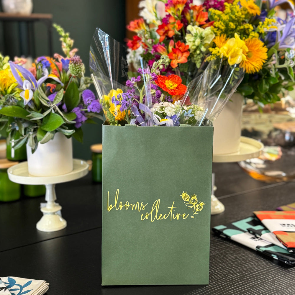 Floral arrangement in a green bag with 'blooms collective' branding on a table with more floral arrangements.
