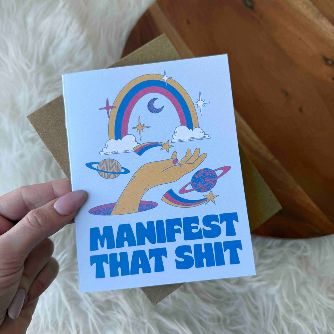 A hand holding a greeting card with a hand-drawn design, featuring a rainbow, clouds, and celestial bodies with the text 'MANIFEST THAT SH*T'.