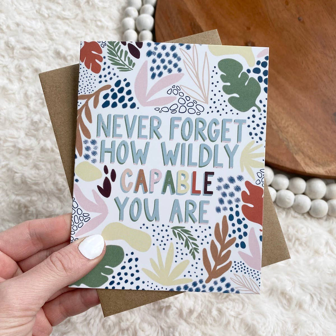 A hand holding a greeting card with a floral pattern and the text 'NEVER FORGET HOW WILDLY CAPABLE YOU ARE' printed on it.