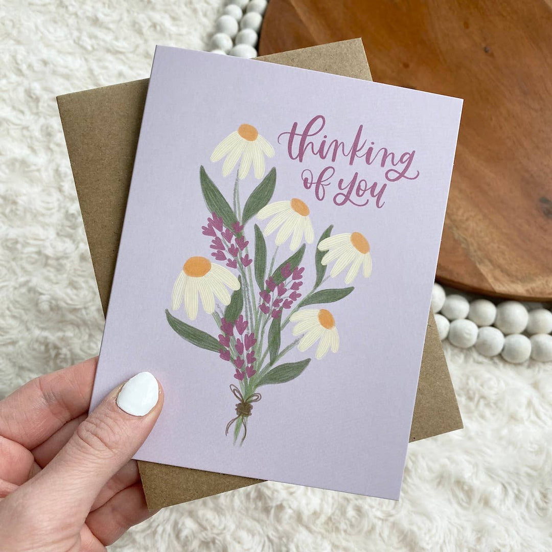 A hand holding a greeting card with a floral design and the text 'thinking of you' on it, paired with a brown envelope.