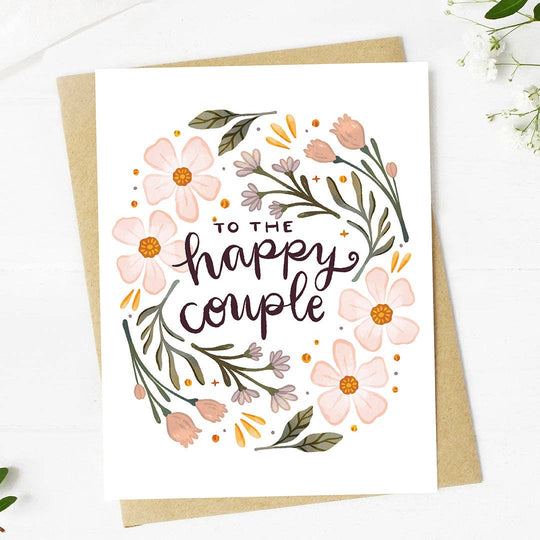 A wedding card with pink floral illustrations and cursive text that reads 'To the happy couple', accompanied by a brown kraft envelope.