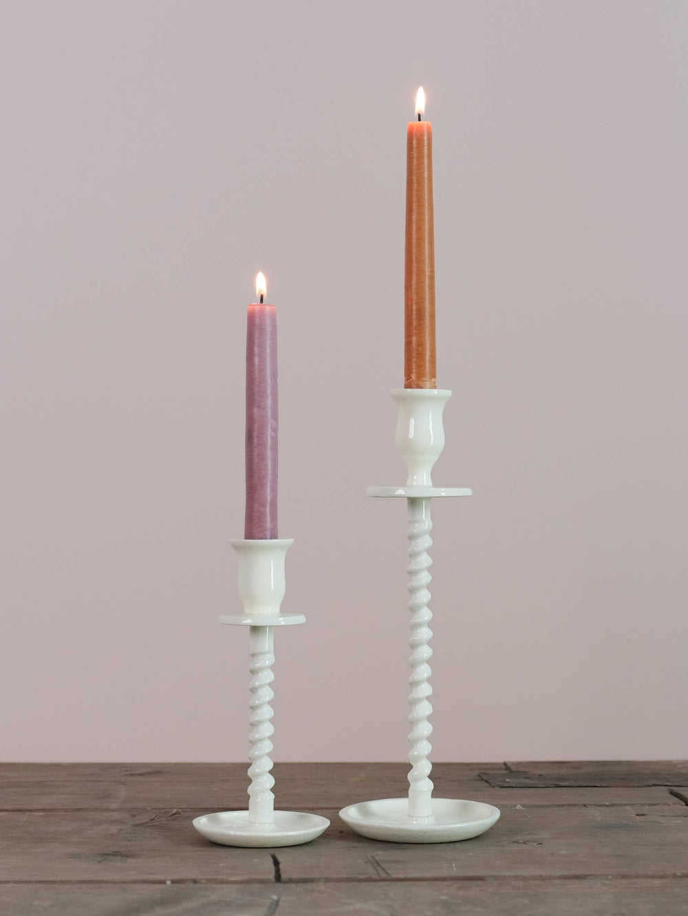 Two candle holders with a corkscrew design, one large in off-white and one small in white, both with lit candles on top, placed on a wooden surface against a neutral background.