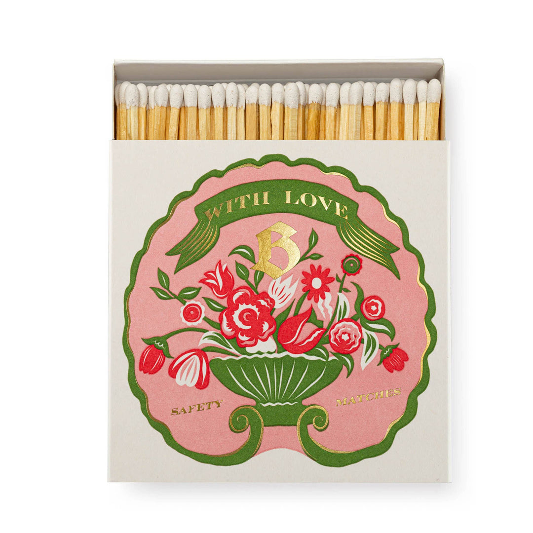 A matchbox with a illustration of a of a floral arrangement in reds, pink and white with With Love Safety Match in gold foil on a pink background.