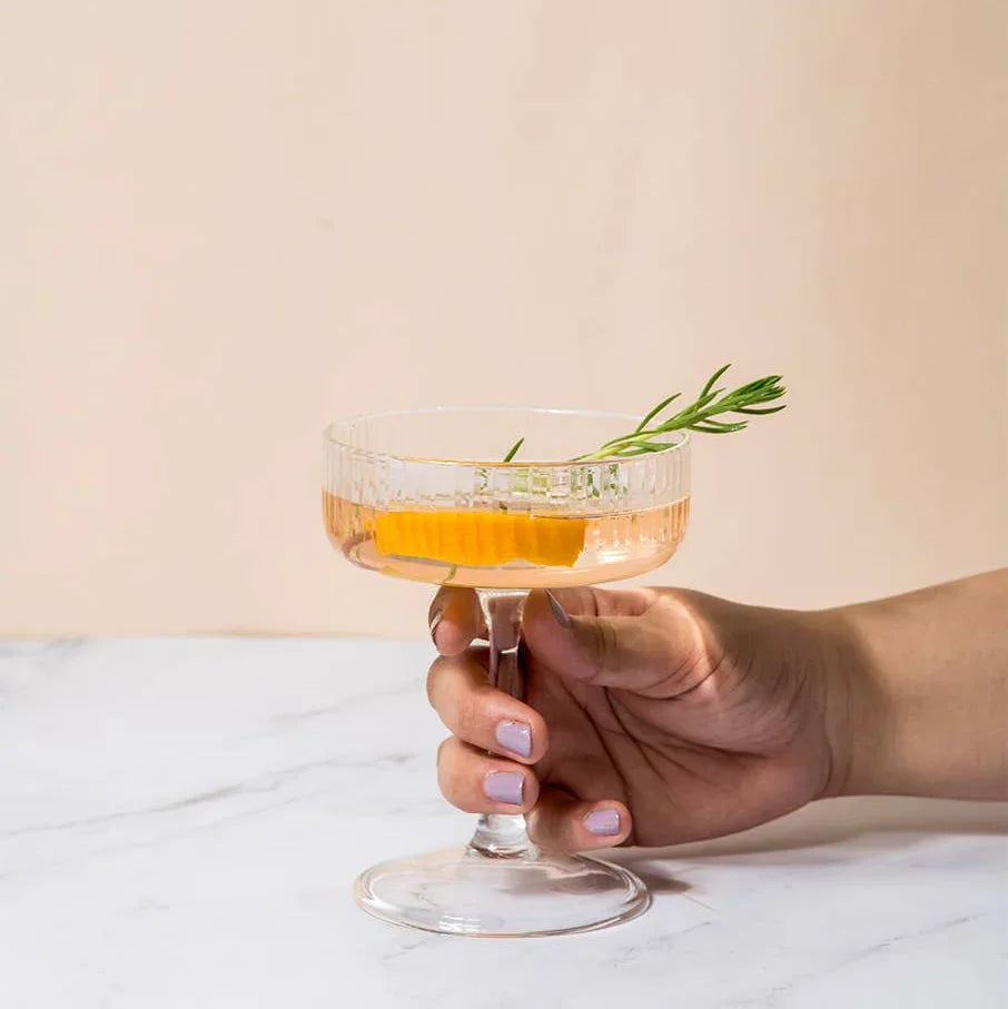 Hand holding cocktail glass with orange slice and rosemary garnish on marble surface