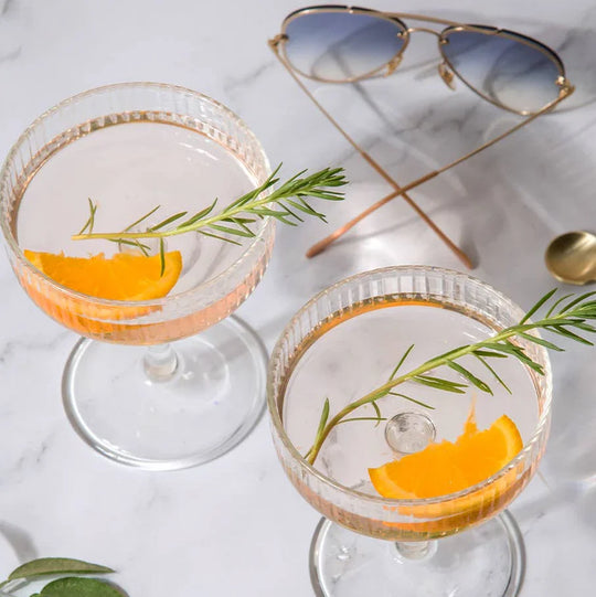 Two elegant cocktails with orange slices and rosemary garnish on marble, styled with sunglasses.