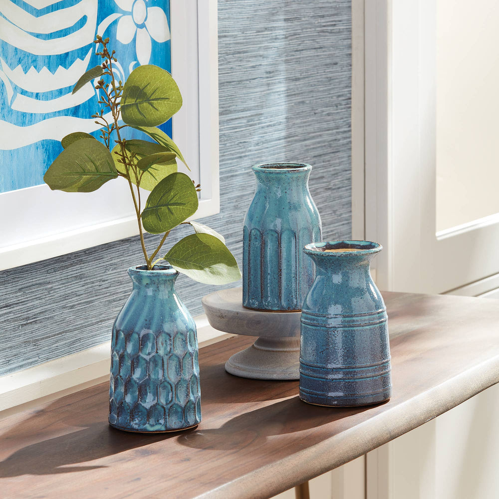 Three blue textured vases on a wooden surface with a decorative painting in the background.