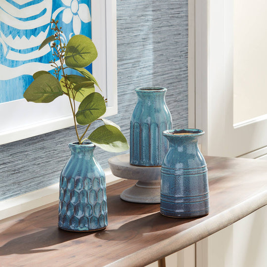 Three blue textured vases on a wooden surface with a decorative painting in the background.