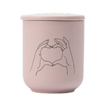 Pink ceramic jar with lid featuring a minimalist line drawing of hands holding a heart on a white background.