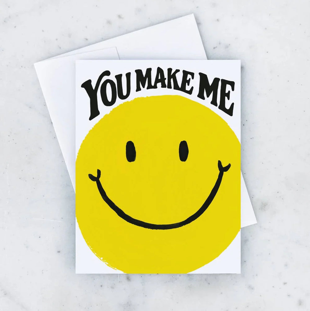 Card with yellow smiley face and 'You Make Me' text on a marble surface