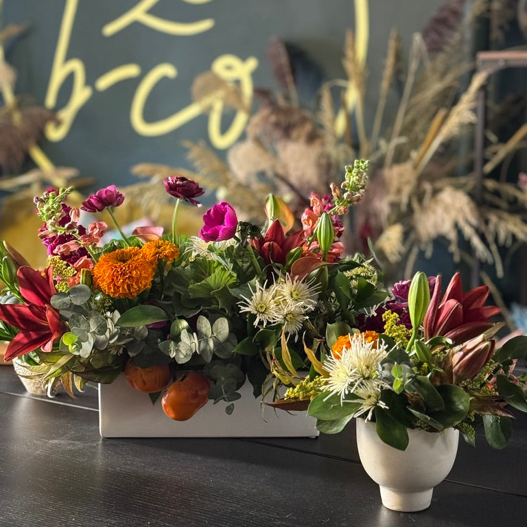 A vibrant floral arrangement featuring red, orange, pink, and white flowers with green foliage, styled in a white rectangular planter and a small white ceramic vase on a dark wooden table.