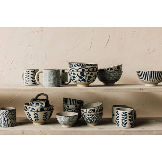 Collection of black and white ceramic bowls and mugs on a wooden shelf against a beige wall.