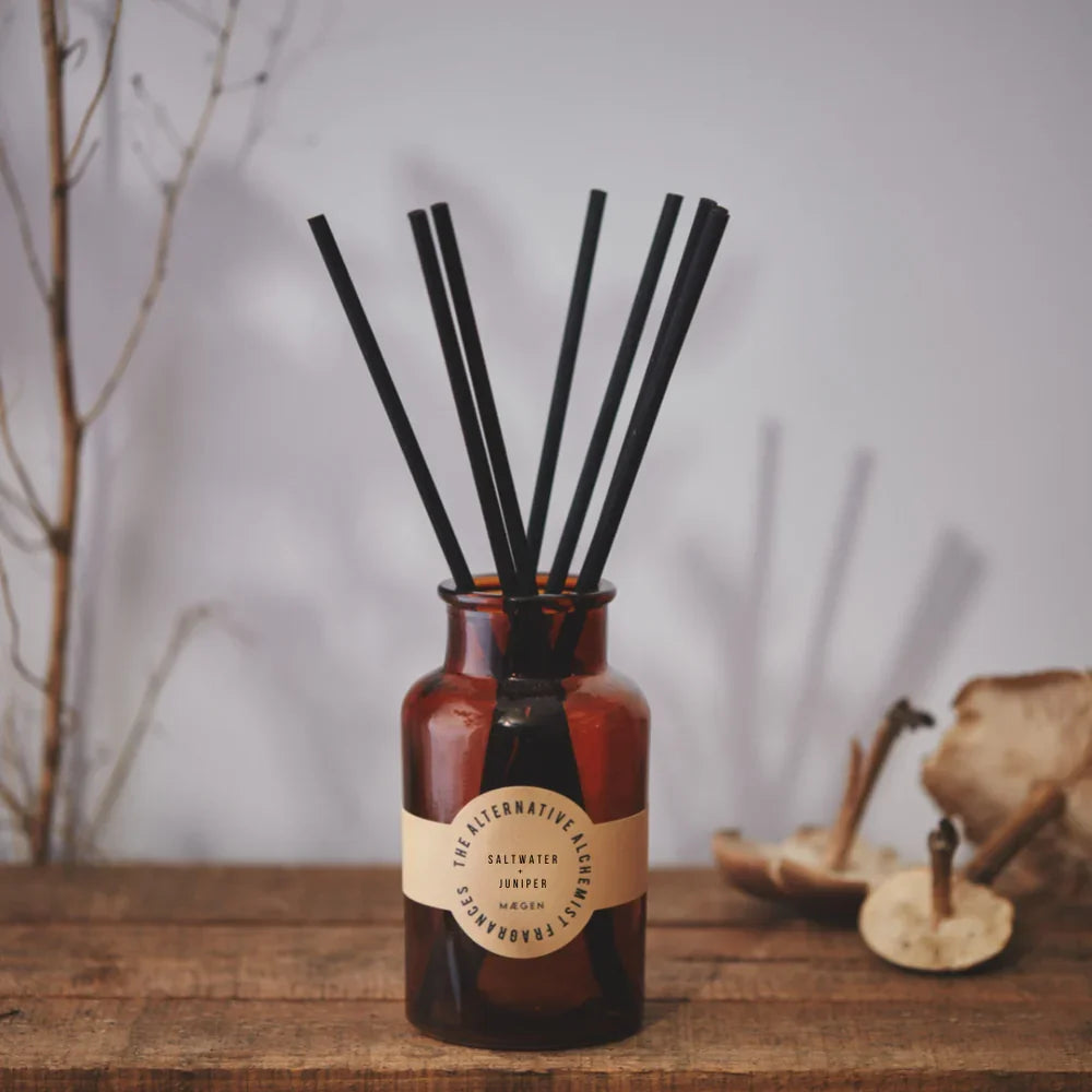 Amber glass reed diffuser with black sticks, labeled Saltwater Juniper, on rustic wood surface