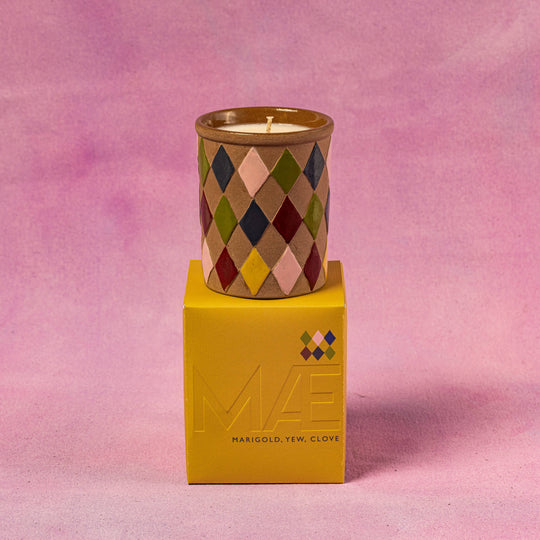 Candle in a patterned container on top of a yellow box with a pink background