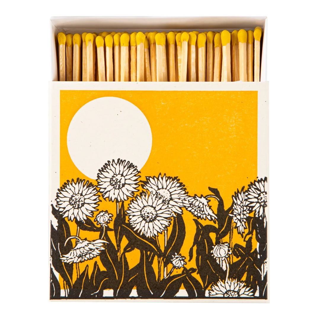 A matchbox package shown open with wooden matches topped in yellow, set against a white background. The front of the box features a graphic design with a large white sun on a mustard-yellow field and black-and-white flower silhouettes along the bottom.