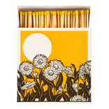 A matchbox package shown open with wooden matches topped in yellow, set against a white background. The front of the box features a graphic design with a large white sun on a mustard-yellow field and black-and-white flower silhouettes along the bottom.