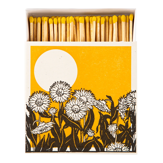 A matchbox package shown open with wooden matches topped in yellow, set against a white background. The front of the box features a graphic design with a large white sun on a mustard-yellow field and black-and-white flower silhouettes along the bottom.