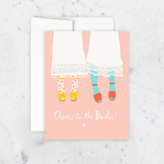 Greeting card with illustration of two women in dresses and colorful shoes on a pink background, with text 'Cheers to the Brides!' on a marble surface.