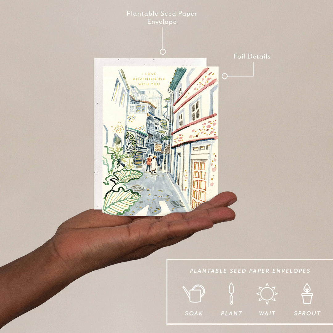 Hand holding a plantable seed paper envelope with a street scene illustration on a beige background.