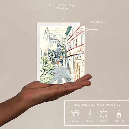 Hand holding a plantable seed paper envelope with a street scene illustration on a beige background.