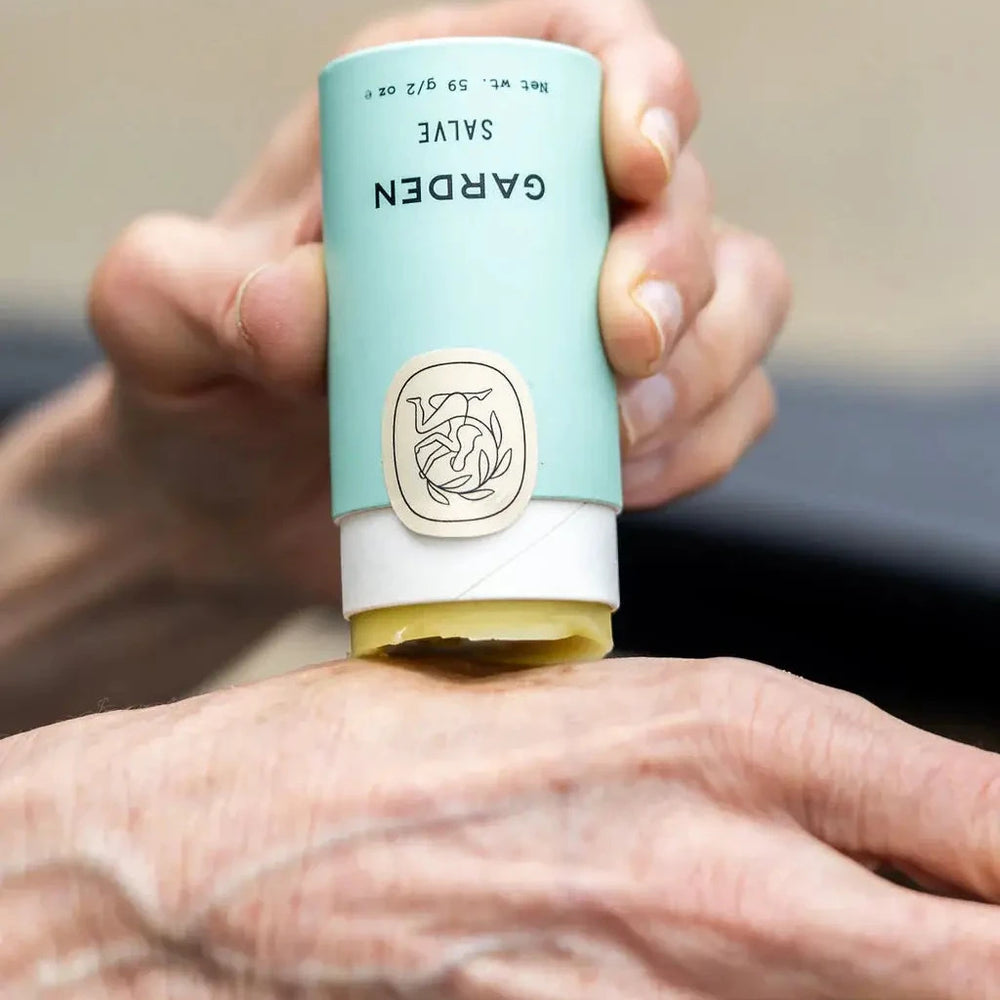 Hand applying salve from a blue and white tube labeled 'Garden' with a logo.