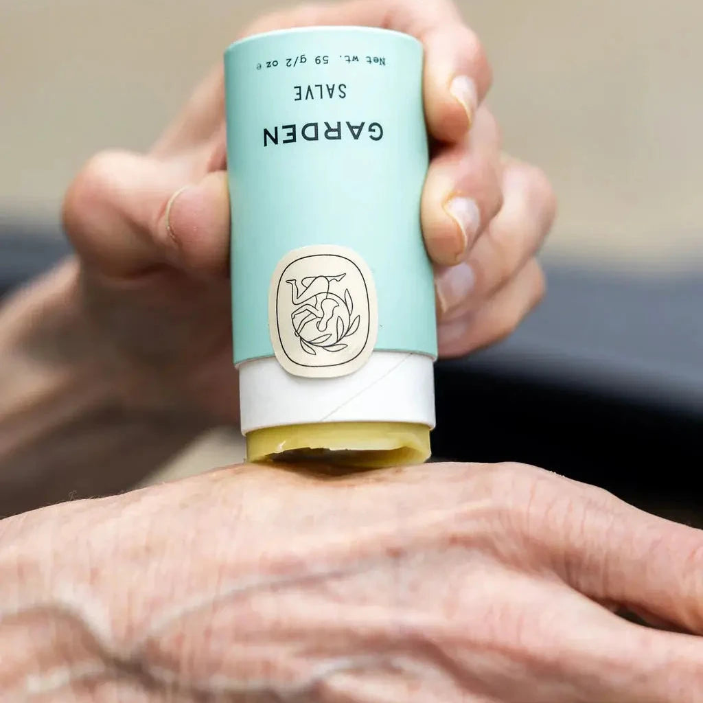 Hand applying salve from a blue and white tube labeled 'Garden' with a logo.