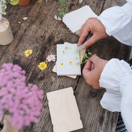 Person arranging flowers on small cards on a wooden table with plants around
