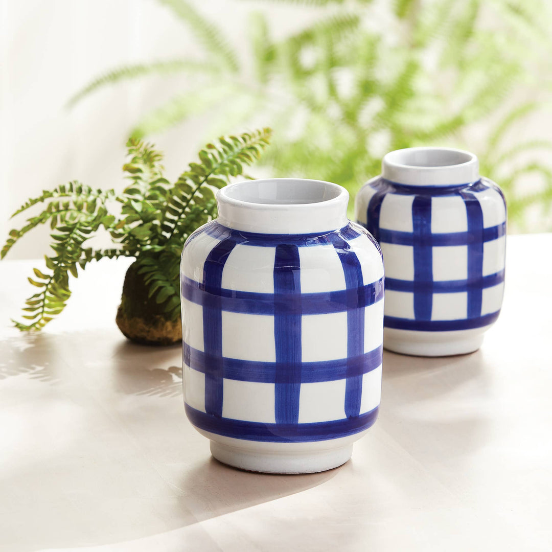 Two ceramic vases with blue and white checkered pattern on a light surface with green plants in the background.