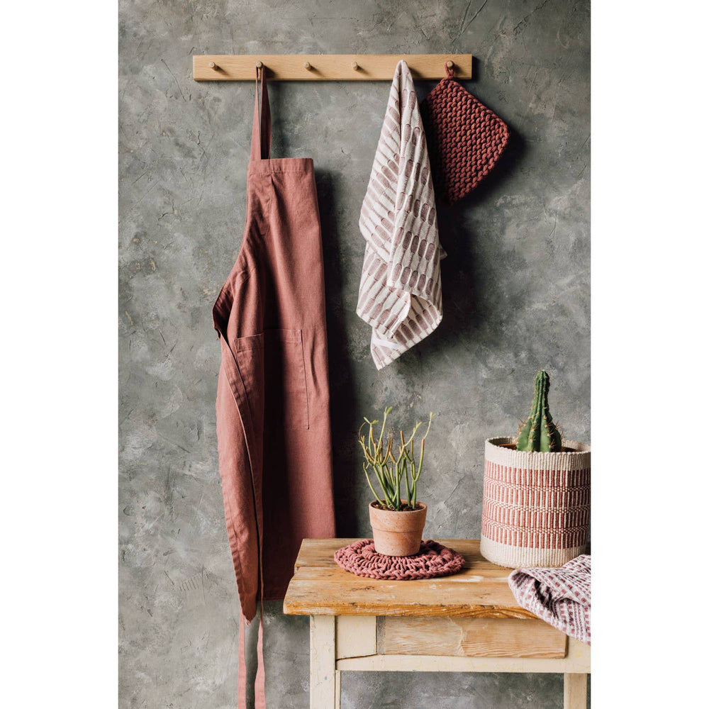 Brown apron, towel, and pot holder hanging on a wooden rack against a gray wall with a small table and plants.