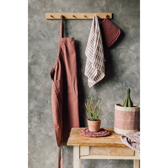 Brown apron, towel, and pot holder hanging on a wooden rack against a gray wall with a small table and plants.