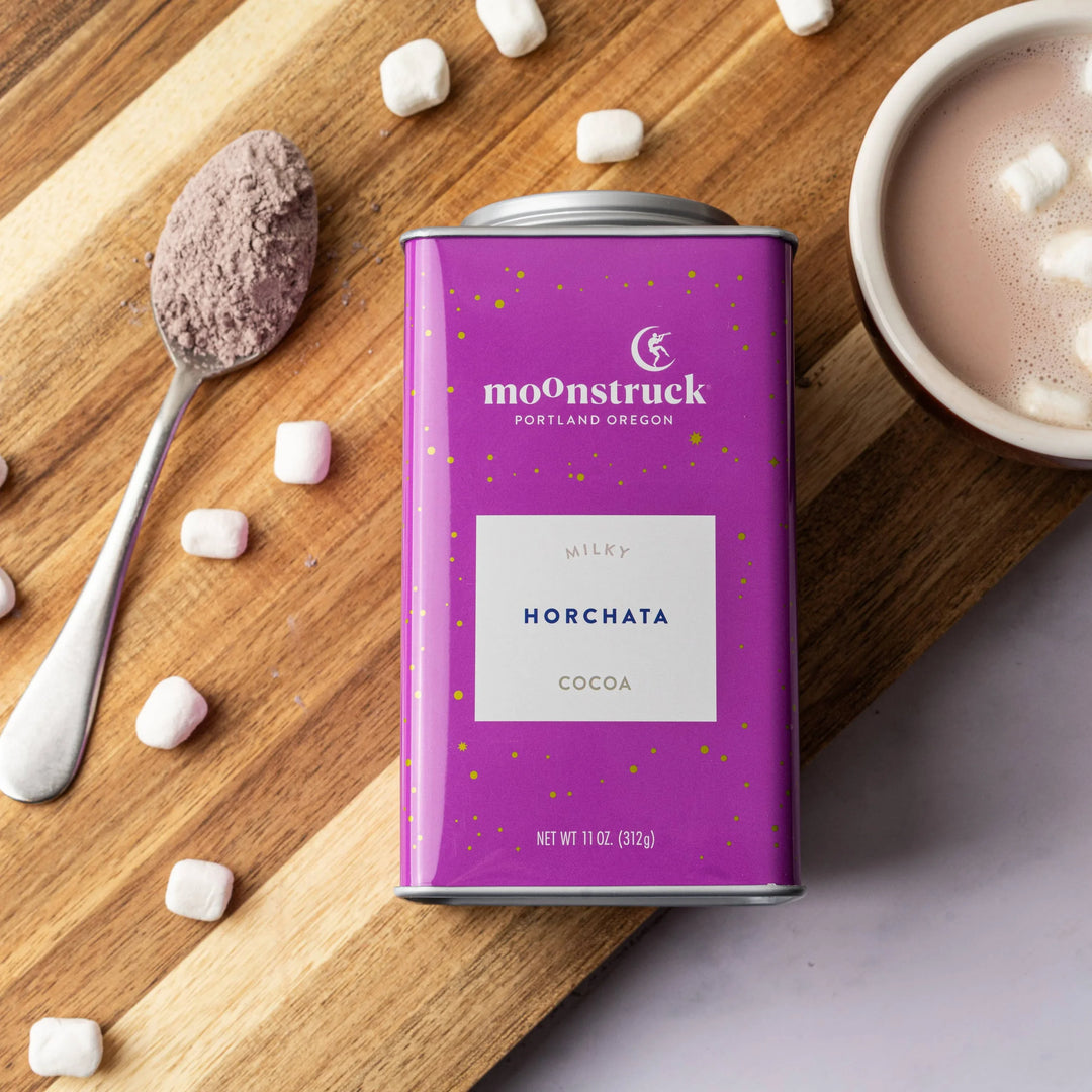 Moonstruck horchata cocoa can on a wooden board with hot chocolate and marshmallows.