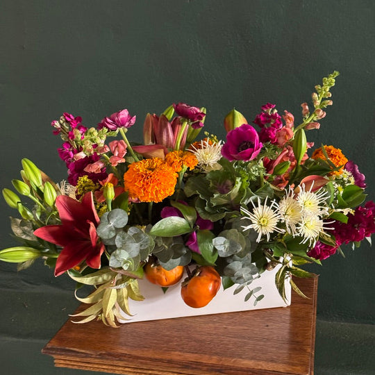 A vibrant, multicolor floral arrangement in a white rectangular planter box, styled on a rustic wooden stool against a dark green wall.