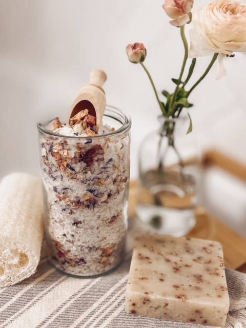 Glass jar with bath salts, wooden spoon, and bar of soap on a striped towel with flowers in the background.